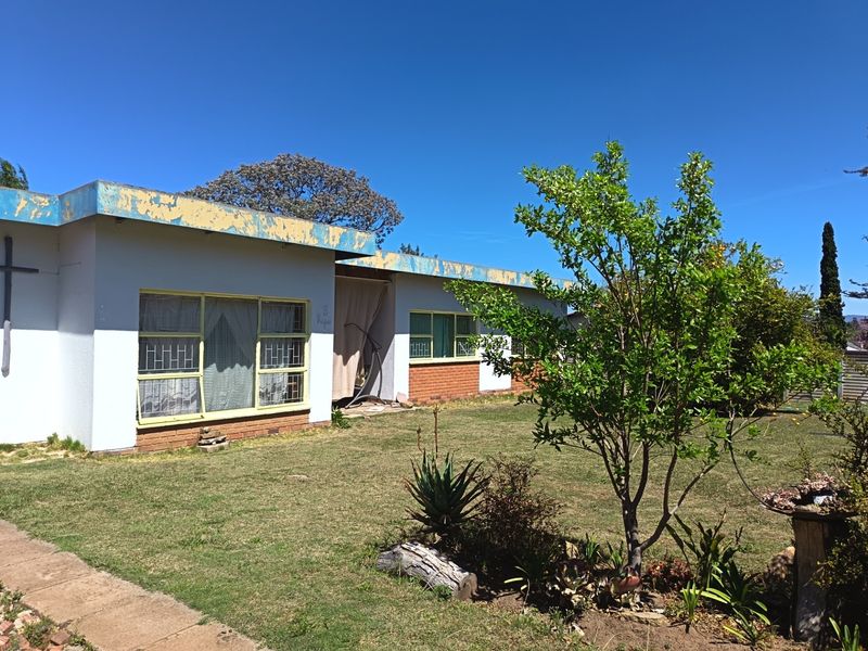 Rear view of a single-story house featuring a brick and plaster facade, security bars on windows, and a lawn area requiring maintenance.