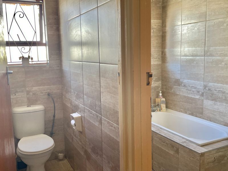 A functional bathroom featuring neutral wood-look wall tiles, a standard bathtub, and a toilet, with natural light provided by a window with security bars.