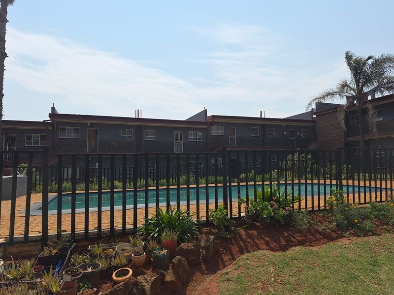 A communal swimming pool area featuring a paved deck, metal security fencing, and established garden beds within an apartment complex.