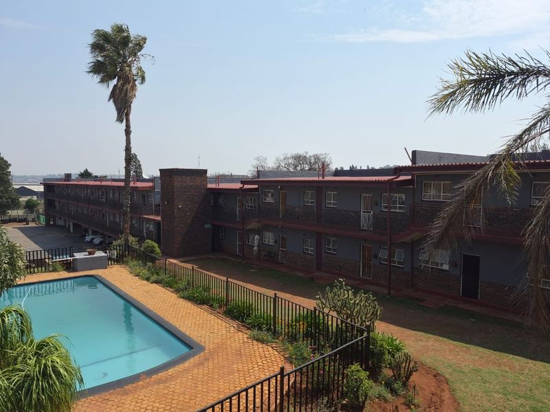 Exterior view of an apartment complex featuring a swimming pool, paved pool area, and multi-story brick building with metal roofing.
