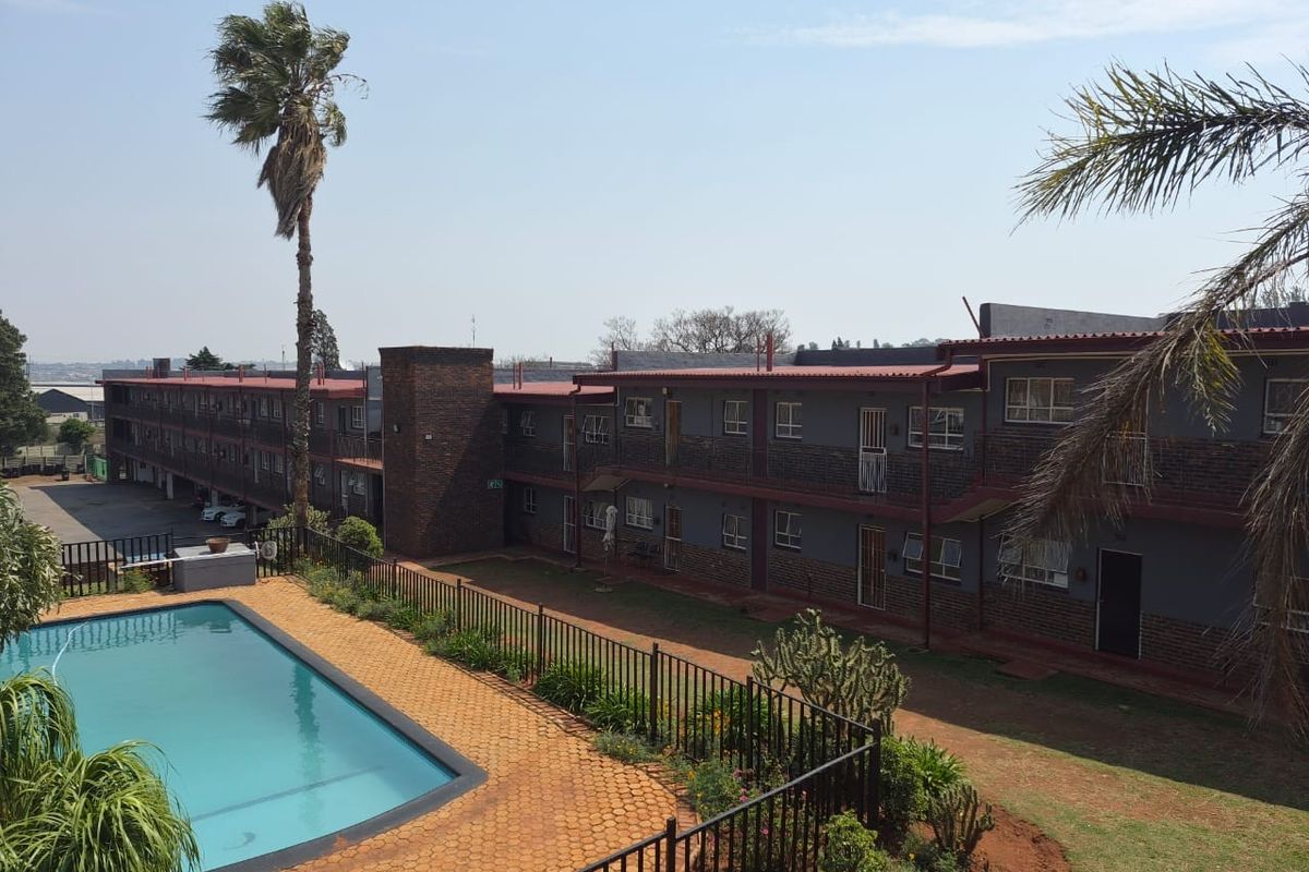 Exterior view of an apartment complex featuring a swimming pool, paved pool area, and multi-story brick building with metal roofing.