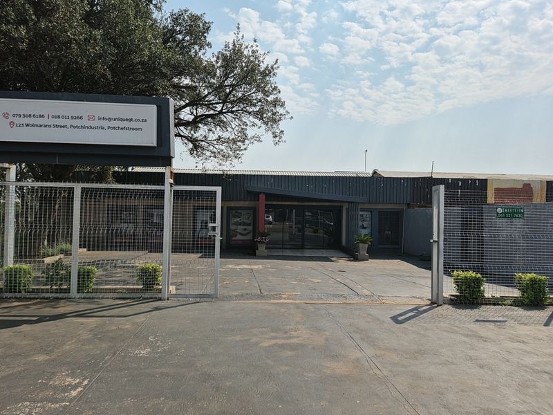 Front view of an industrial property featuring a paved driveway, secure metal fencing, and prominent commercial signage at the entrance.