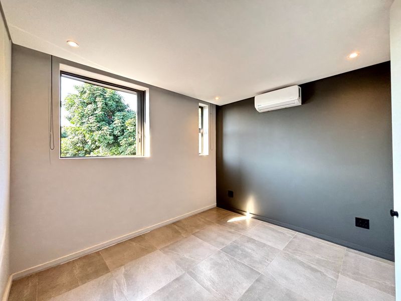 A bright, modern, and empty bedroom featuring neutral tiled flooring, recessed lighting, and a wall-mounted air conditioning unit.