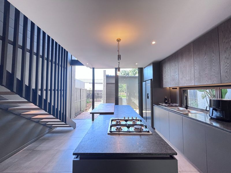 Modern kitchen featuring a central island with gas cooktop, sleek cabinetry, and an adjacent staircase, opening onto a rear patio area.