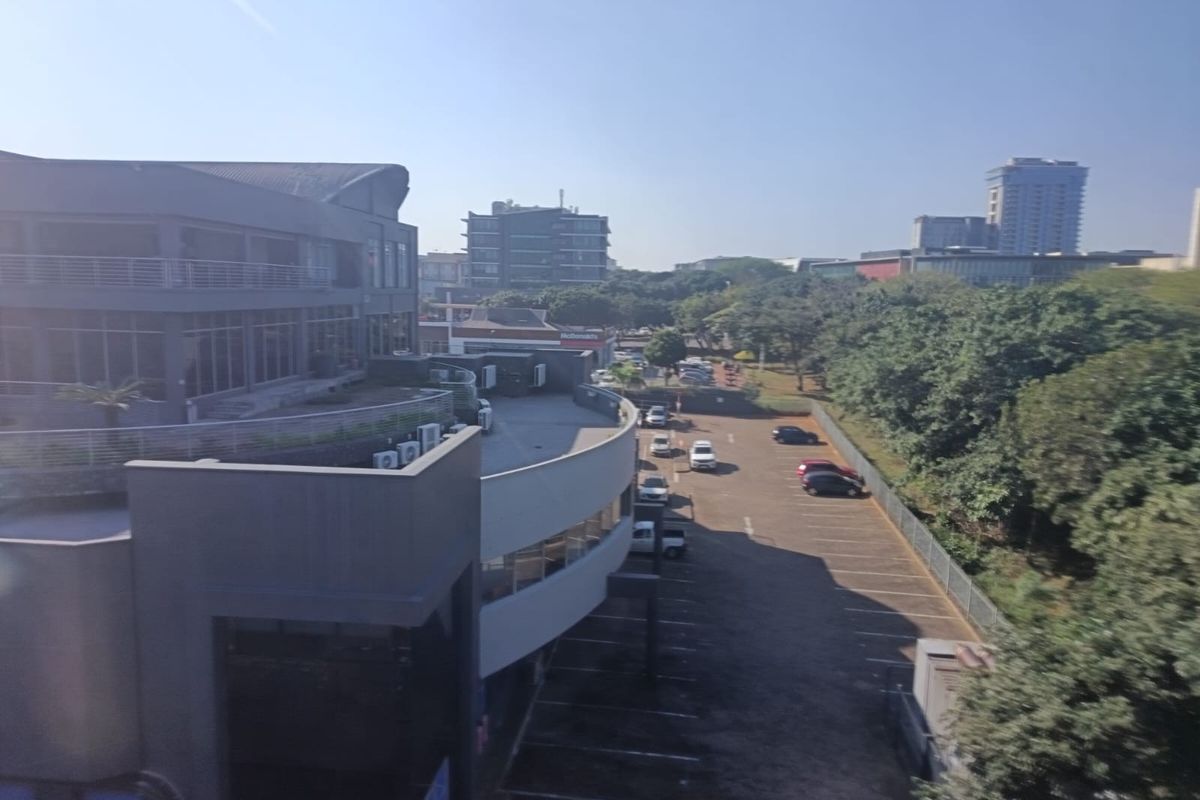 Aerial view of the office building exterior, featuring a paved parking area, modern architectural facade, and surrounding landscaped grounds.