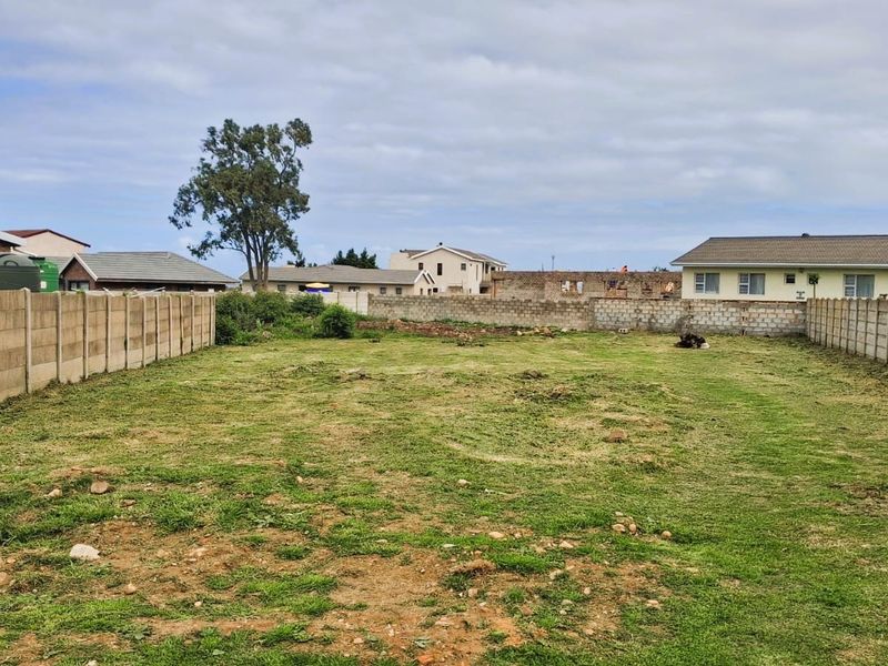 A vacant residential plot enclosed by precast concrete boundary walls, featuring a grassed surface and surrounding neighborhood views.