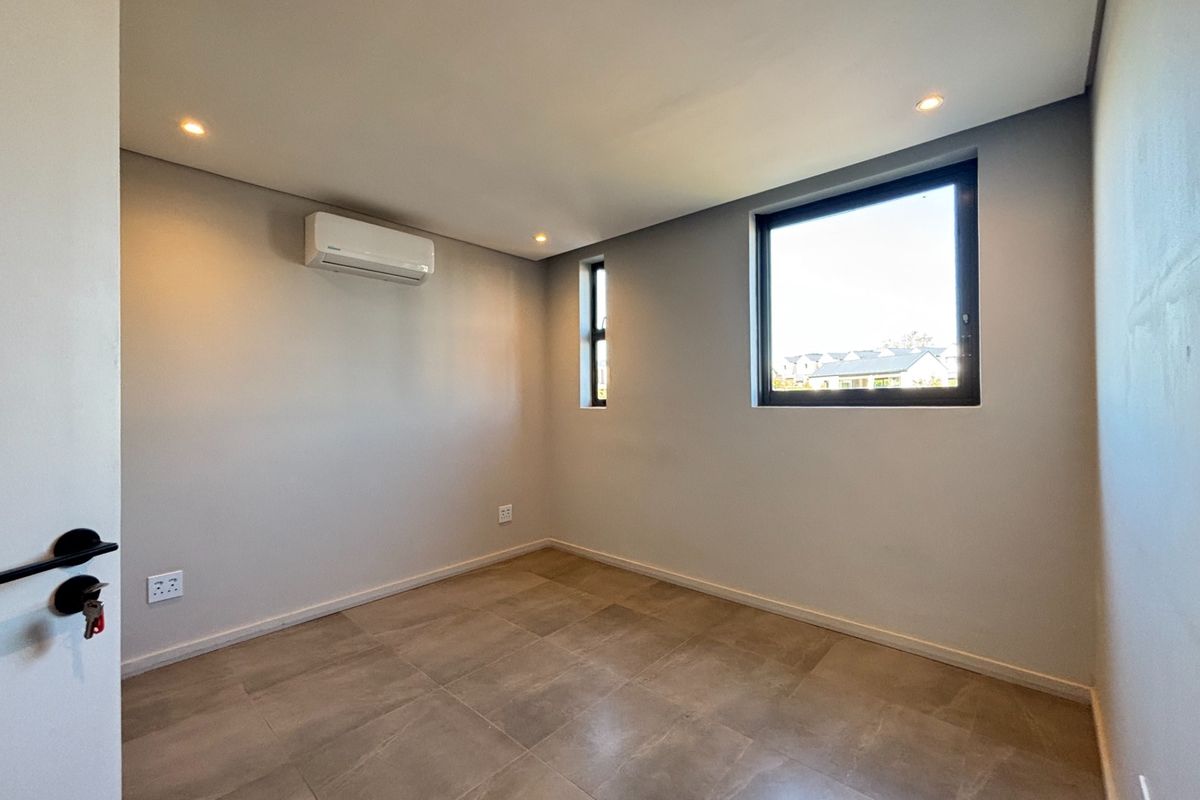 A bright, modern, and empty bedroom featuring neutral tiled flooring, recessed downlighting, and a wall-mounted air conditioning unit.