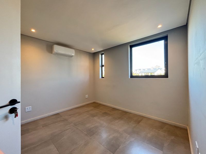 A bright, modern, and empty bedroom featuring neutral tiled flooring, recessed downlighting, and a wall-mounted air conditioning unit.