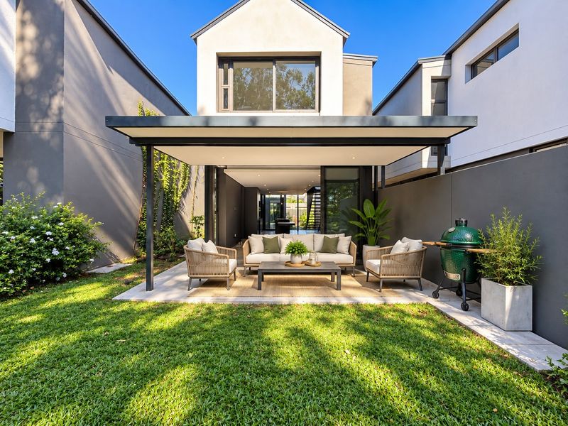 A modern rear townhouse exterior featuring a covered patio, paved terrace, and a well-maintained garden, seamlessly connecting to the interior living space.