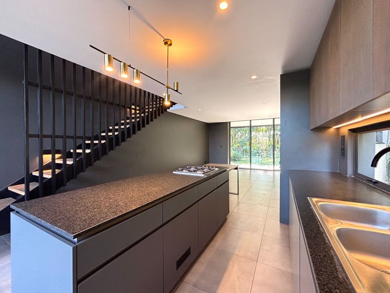 Modern open-plan kitchen featuring a central island with gas cooktop, sleek cabinetry, and a striking staircase, leading to a bright living area with garden access.