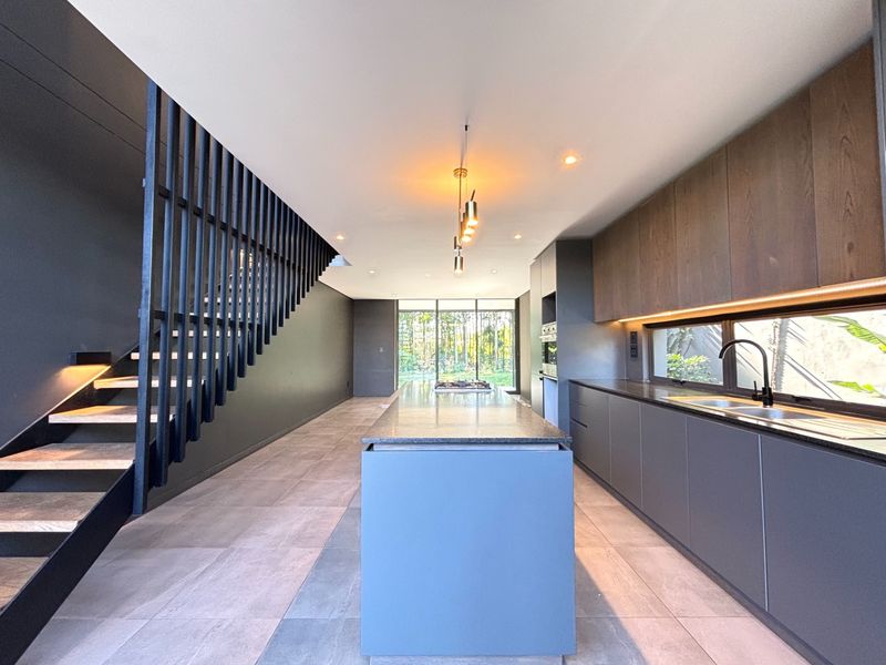 Modern open-plan kitchen featuring a central island, sleek dark cabinetry, integrated appliances, and a striking staircase, leading to a bright rear garden view.