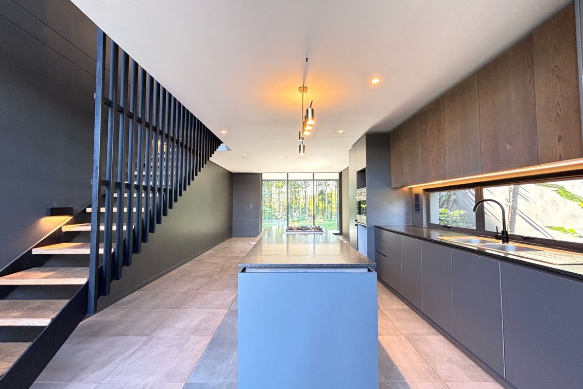 Modern open-plan kitchen featuring a central island, sleek dark cabinetry, integrated appliances, and a striking staircase, leading to a bright rear garden view.