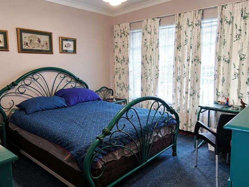 A standard bedroom featuring neutral wall tones, carpeted flooring, and large windows fitted with venetian blinds and curtains.