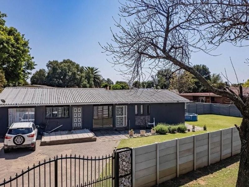 Front exterior view of a single-story house featuring a corrugated metal roof, paved driveway, and secure perimeter walling with a wrought iron gate.