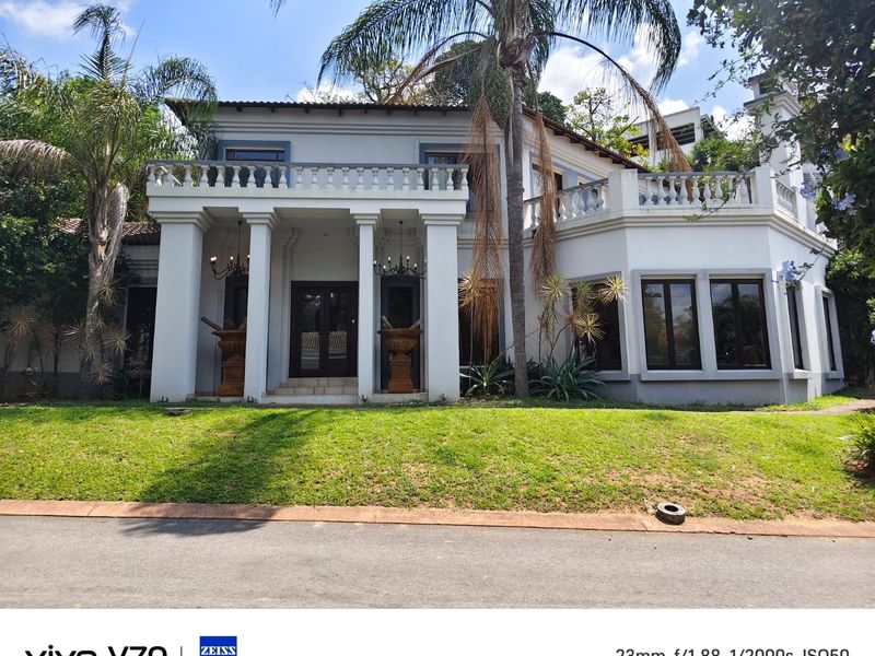 Front elevation of a two-story residence featuring a prominent columned entrance, upper-level terrace with balustrade, and mature landscaping.