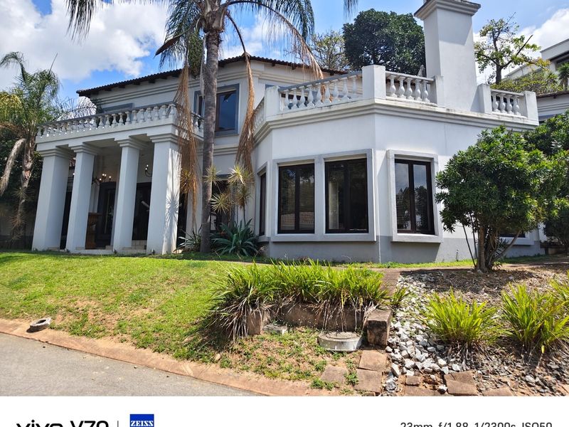 A classic two-story residence featuring a prominent front terrace with decorative balustrades, structural columns, and a chimney, set within a landscaped garden.