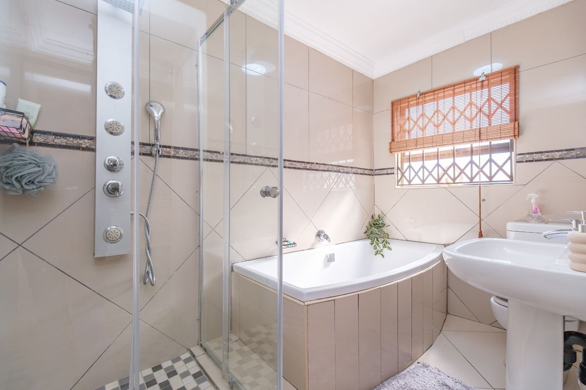 A well-maintained bathroom featuring a glass-enclosed shower with a modern shower panel, a built-in bathtub, a pedestal sink, and neutral wall tiling.