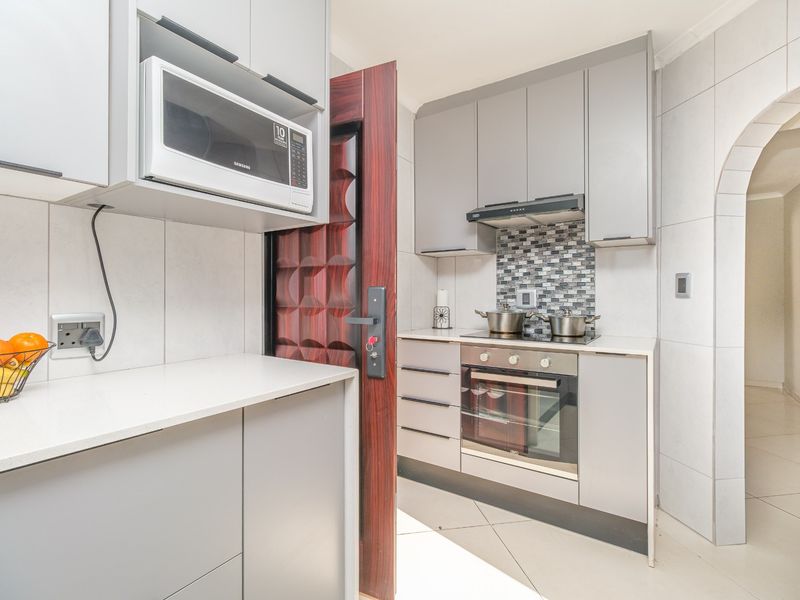 Modern kitchen featuring grey cabinetry, stone countertops, built-in oven, hob, extractor fan, and a tiled backsplash, leading through an arched doorway.