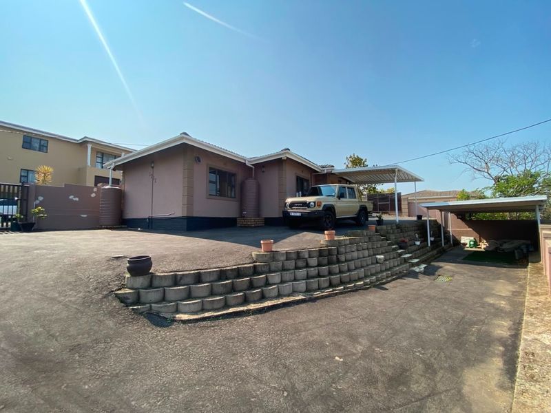 Front exterior view of the house featuring a large asphalt driveway, retaining wall, carport, and water storage tanks.
