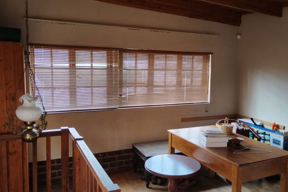 A living area featuring exposed timber ceiling beams, timber flooring, and a wooden balustrade, offering a rustic aesthetic with natural light through venetian blinds.