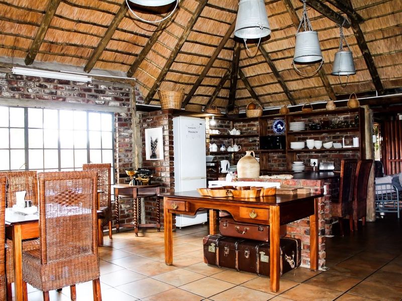 Open-plan living and kitchen area featuring a rustic thatched roof, exposed brick walls, and tiled flooring in a guest house setting.