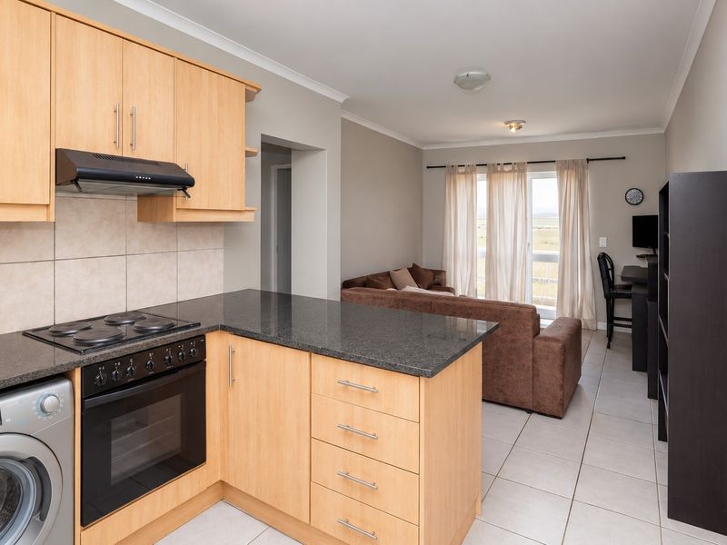 Open-plan kitchen and living area featuring light wood cabinetry, granite countertops, built-in appliances, and tiled flooring with access to a balcony.