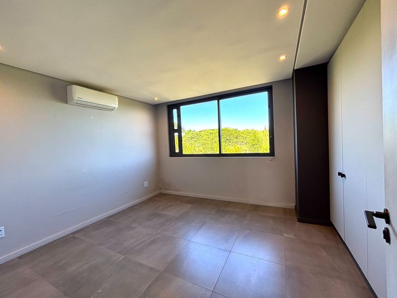 A bright, empty bedroom featuring tiled flooring, built-in cupboards, a split-system air conditioner, and large windows with a pleasant green outlook.
