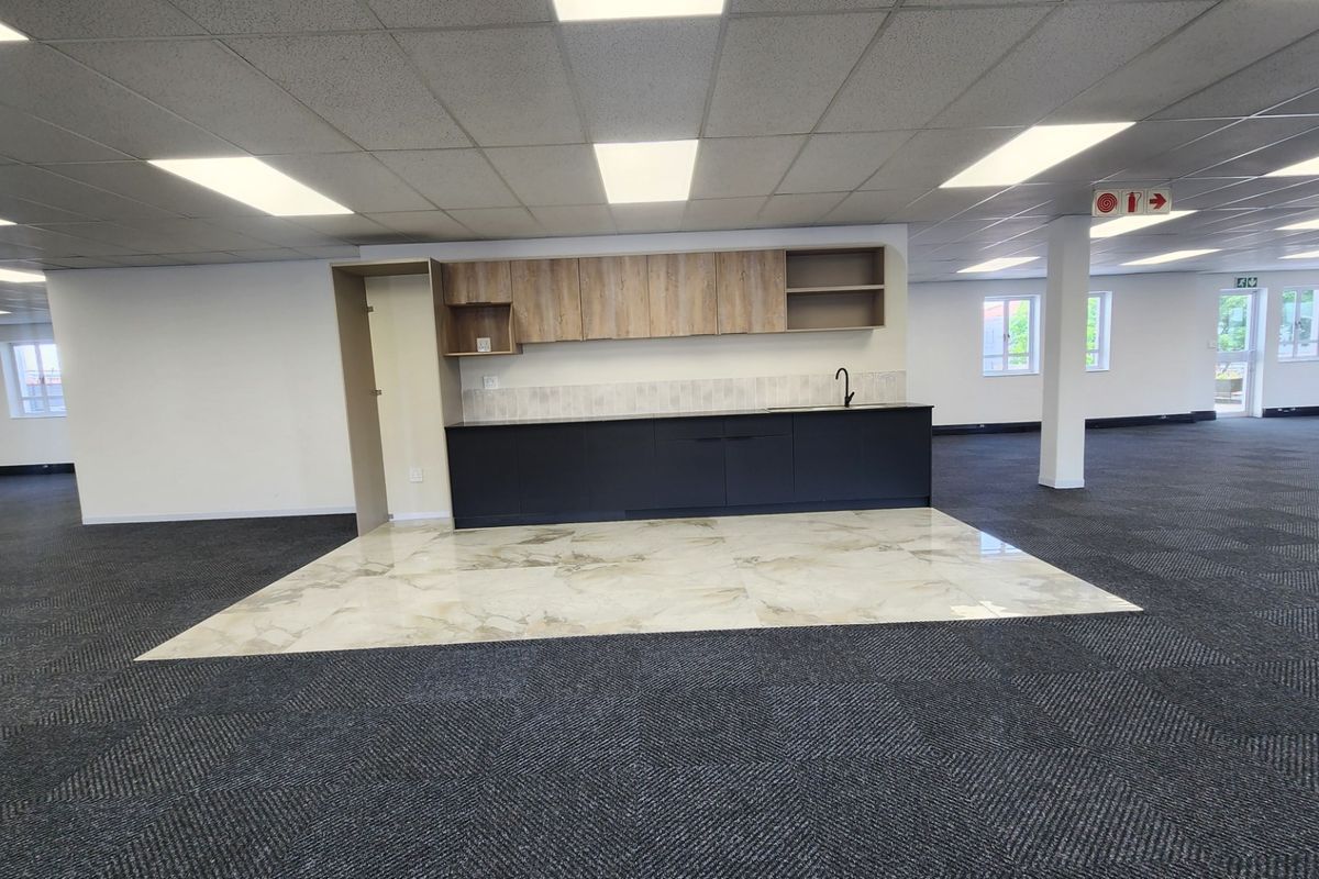 A modern, open-plan office kitchenette featuring dark base cabinets, light wood-look upper cupboards, a sink, and a distinct tiled floor area.