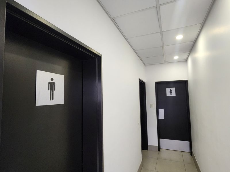 A clean, well-lit commercial hallway featuring tiled flooring, a suspended ceiling, and access doors to male and female restroom facilities.