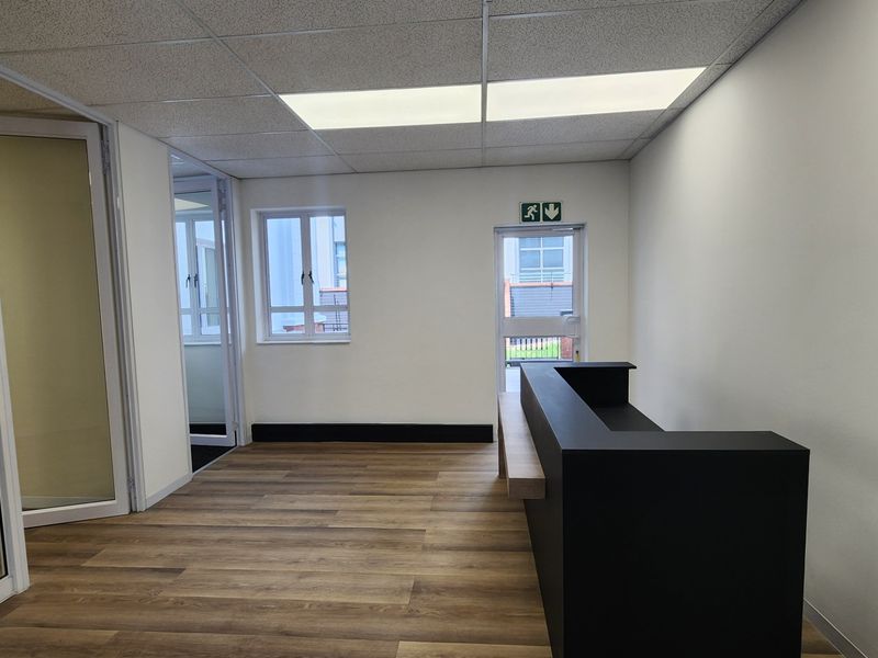 A modern commercial office space featuring a built-in reception desk, wood-look laminate flooring, glass partitions, and a suspended ceiling with recessed lighting.