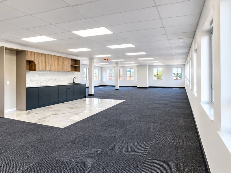 Spacious open-plan commercial office area featuring a modern kitchenette, carpeted flooring, suspended ceilings with recessed lighting, and large windows providing ample natural light.