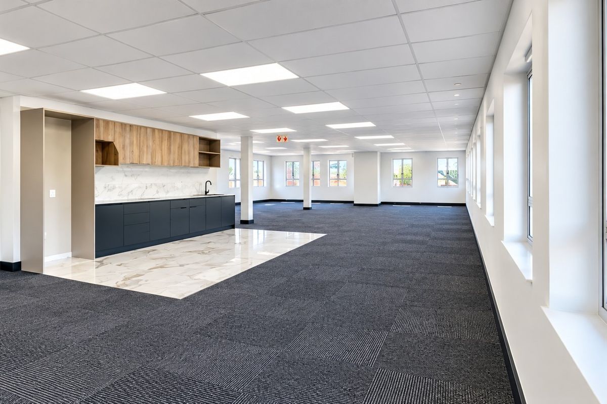 Spacious open-plan commercial office area featuring a modern kitchenette, carpeted flooring, suspended ceilings with recessed lighting, and large windows providing ample natural light.