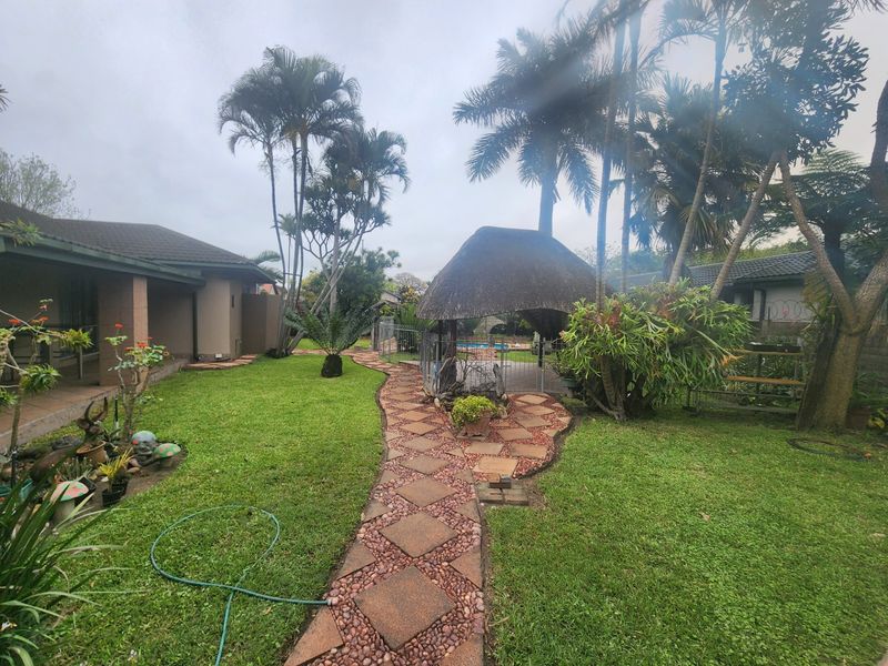 A spacious garden featuring a paved walkway, mature palm trees, and a thatched lapa area overlooking the swimming pool.