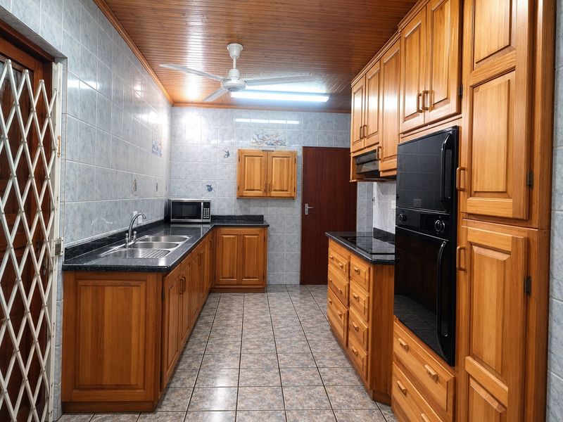 A functional kitchen featuring extensive wooden cabinetry, granite countertops, built-in oven, electric hob, and tiled finishes throughout.