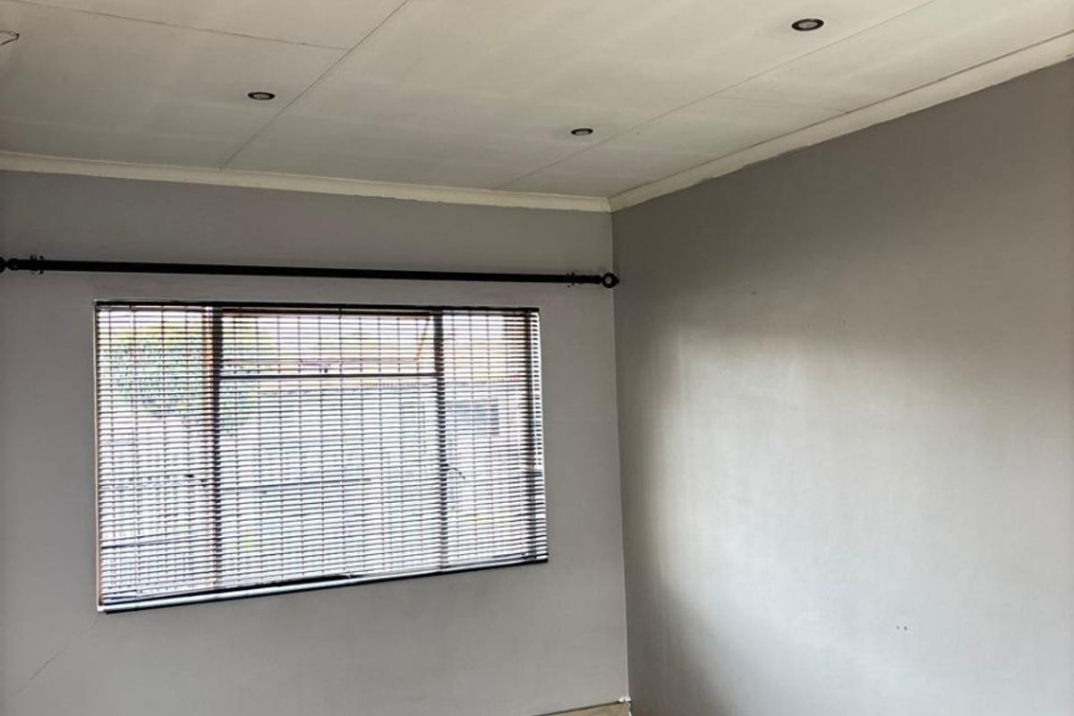 A bright, empty bedroom featuring neutral grey walls, tiled flooring, recessed ceiling lighting, and fitted venetian blinds.