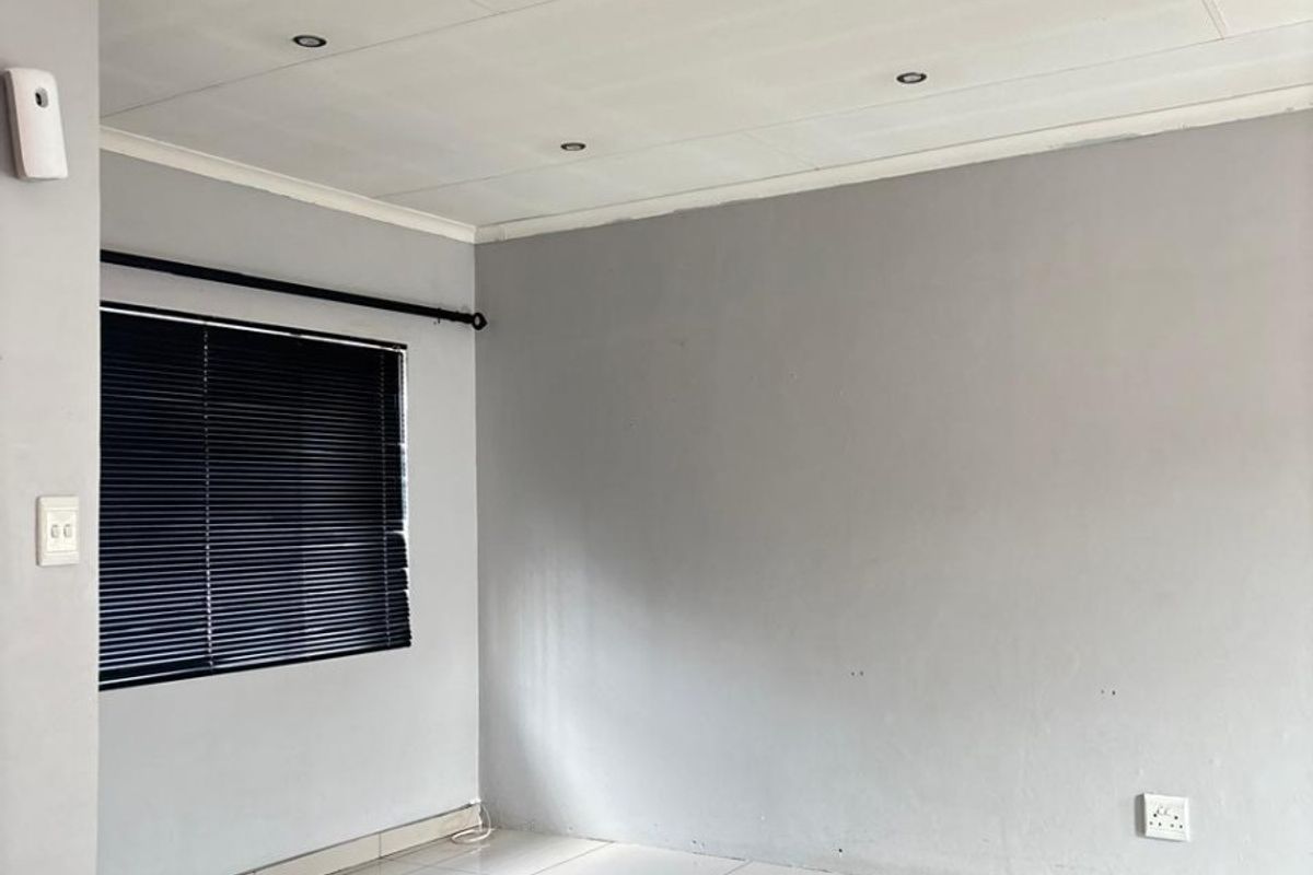 A bright, empty living area featuring neutral grey walls, white tiled flooring, and recessed ceiling lighting with fitted venetian blinds.