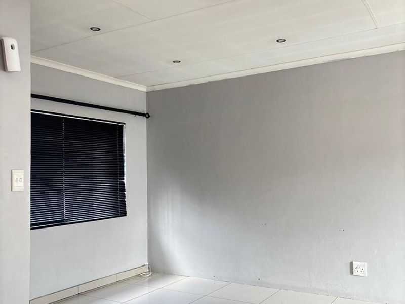 A bright, empty living area featuring neutral grey walls, white tiled flooring, and recessed ceiling lighting with fitted venetian blinds.