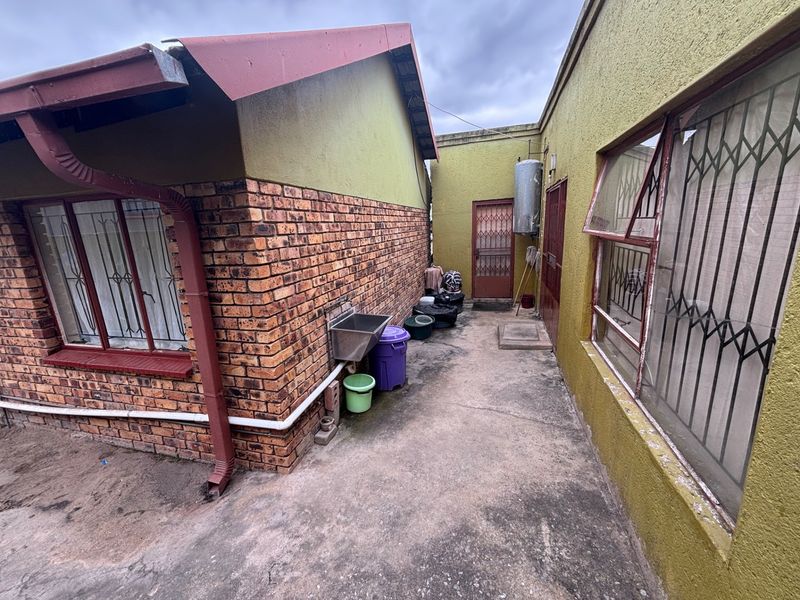 Rear exterior area featuring brick and rendered walls, concrete paving, security gates, and an external wash basin with an attached electric geyser.