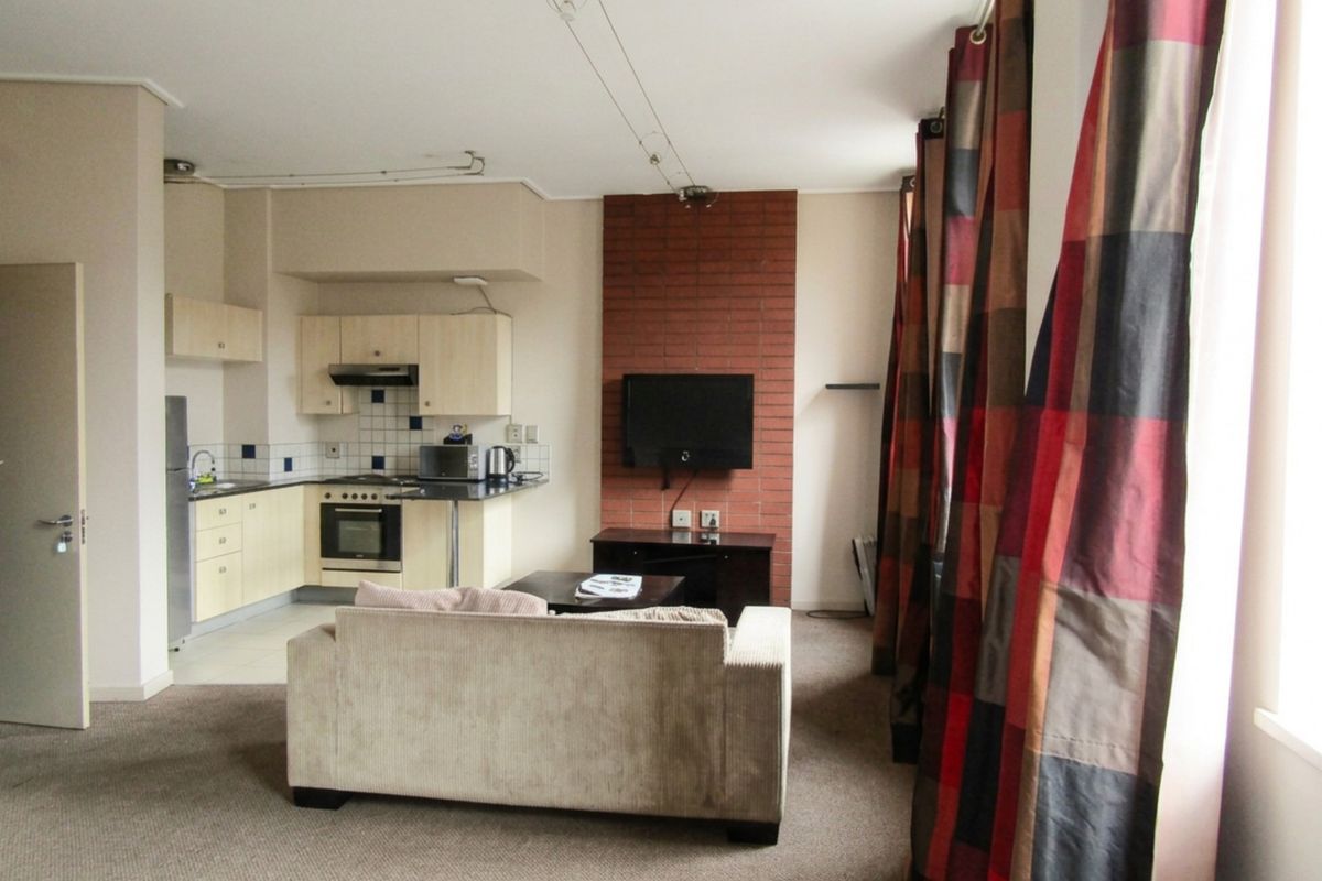 Open-plan living area featuring a compact kitchen with built-in cabinetry and appliances, adjacent to a living space with an exposed brick feature wall.