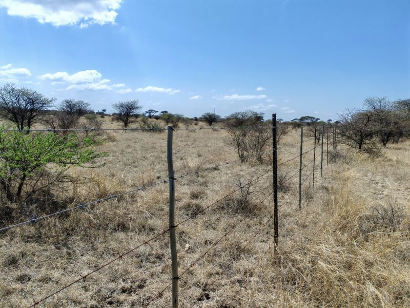A view of a vacant residential plot featuring dry grassland, native shrubs, and perimeter barbed wire fencing with metal and wooden posts.