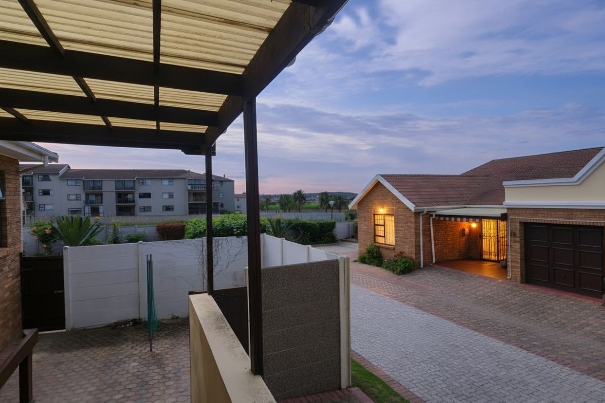 A view of the rear exterior area featuring a paved driveway, a covered patio with a polycarbonate roof, and brick townhouse facades.