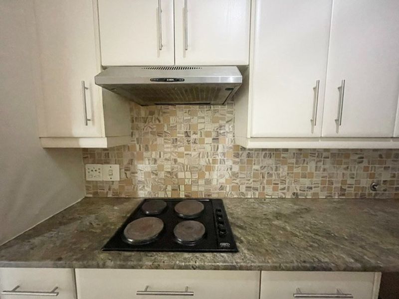 A clean kitchen workspace featuring white cabinetry, a stone countertop, a mosaic tile backsplash, and a built-in electric hob with an extractor fan.