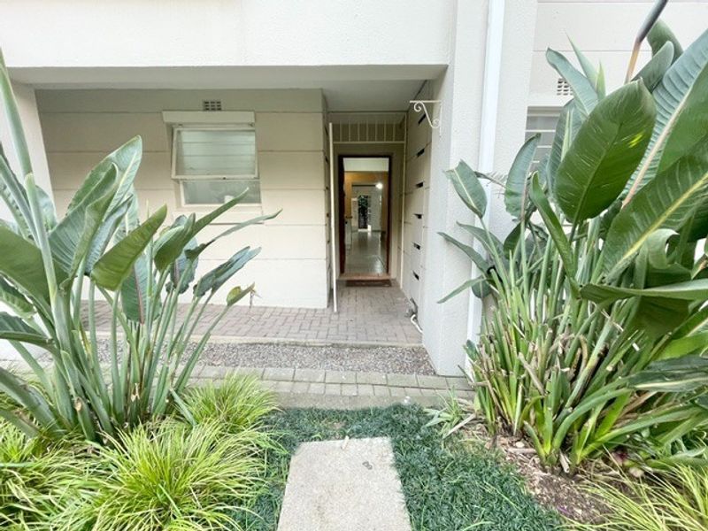 A ground-level apartment entrance featuring a paved walkway, established garden landscaping, and a secure front door access point.