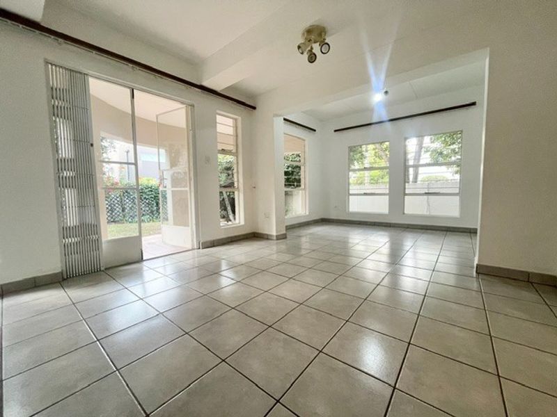 A bright, empty living area featuring neutral ceramic floor tiles, large windows, and sliding glass doors providing access to the outdoor space.