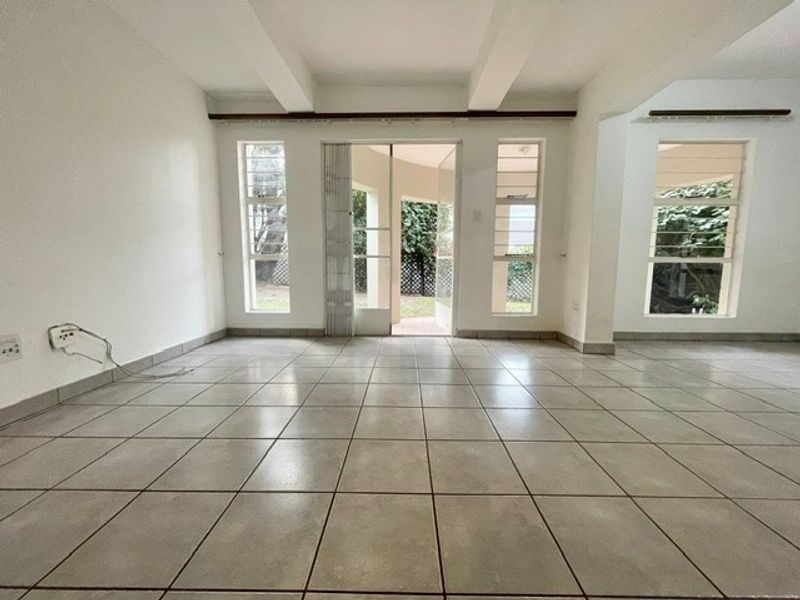 An empty, open-plan living area featuring neutral tiled flooring, exposed ceiling beams, and large windows providing natural light and access to the outdoor space.