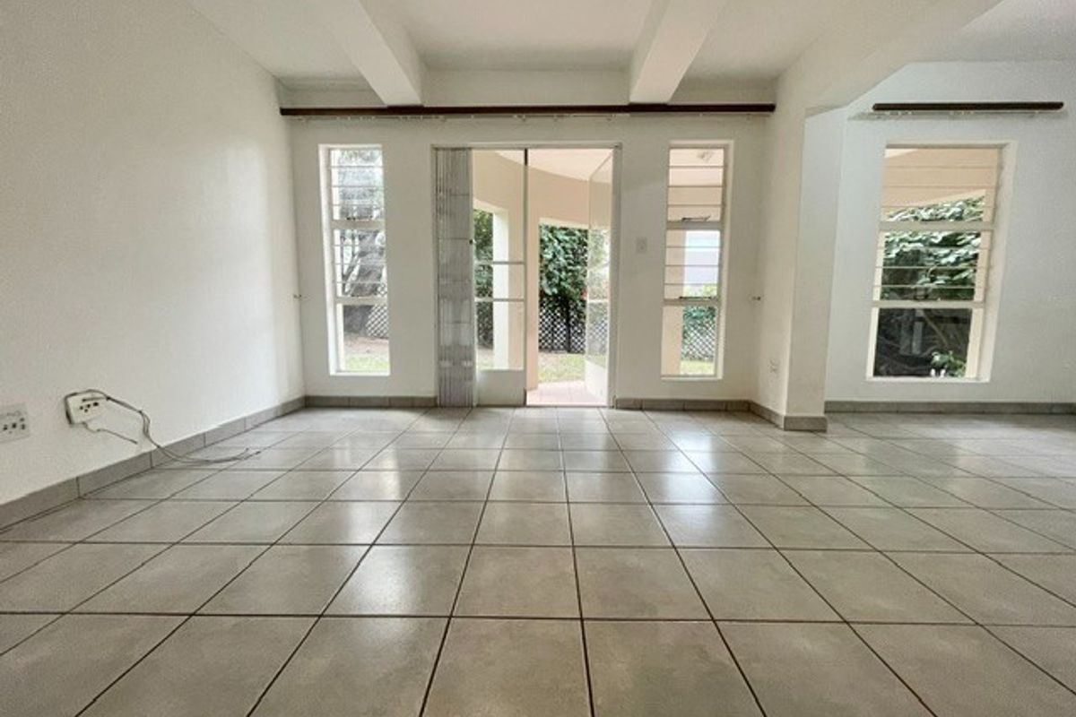 An empty, open-plan living area featuring neutral tiled flooring, exposed ceiling beams, and large windows providing natural light and access to the outdoor space.
