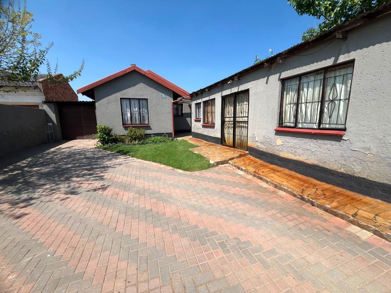 Front exterior view of the property featuring a paved driveway, security gates, and a small garden area with visible exterior wall maintenance required.