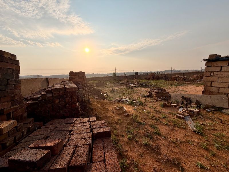 A vacant residential plot featuring a perimeter wall and scattered brick piles, offering a clear view of the surrounding landscape at sunset.