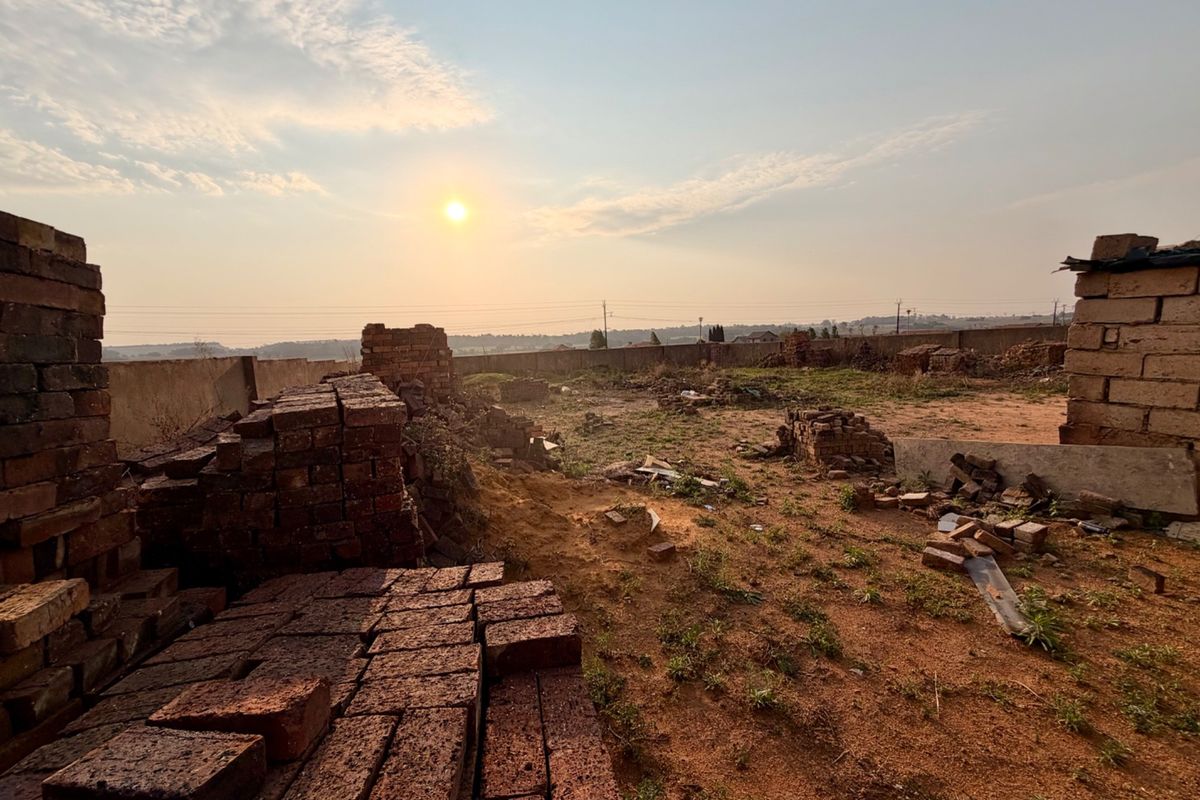 A vacant residential plot featuring a perimeter wall and scattered brick piles, offering a clear view of the surrounding landscape at sunset.