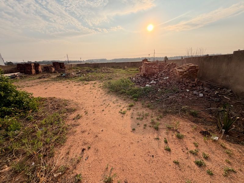 A vacant residential plot featuring a boundary wall, sandy terrain, and scattered construction debris under a sunset sky.